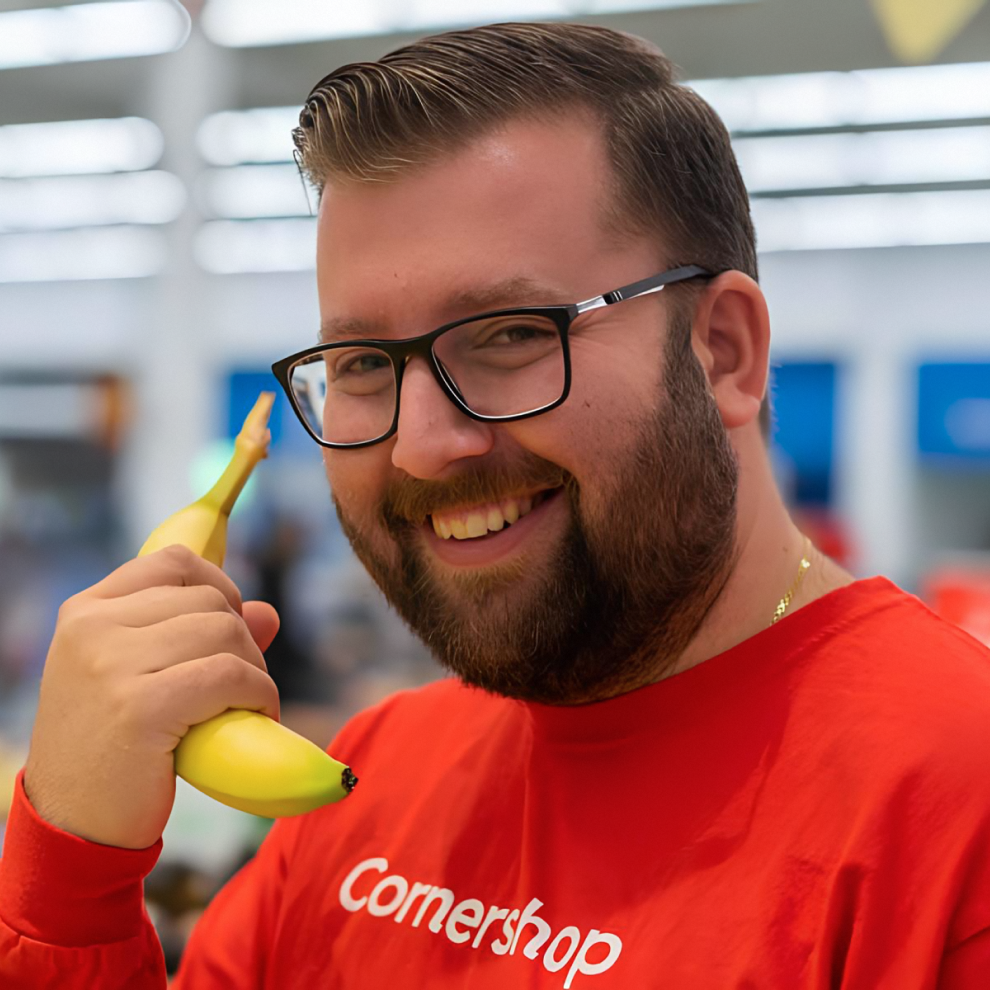 A young man in a red Cornershop shirt smiles while holding a banana to his ear, pretending it's a phone.